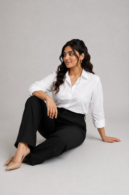 A woman sits on the floor against a light gray studio background, wearing a white button-up shirt by Purple Imli  with rolled sleeves and black trousers.