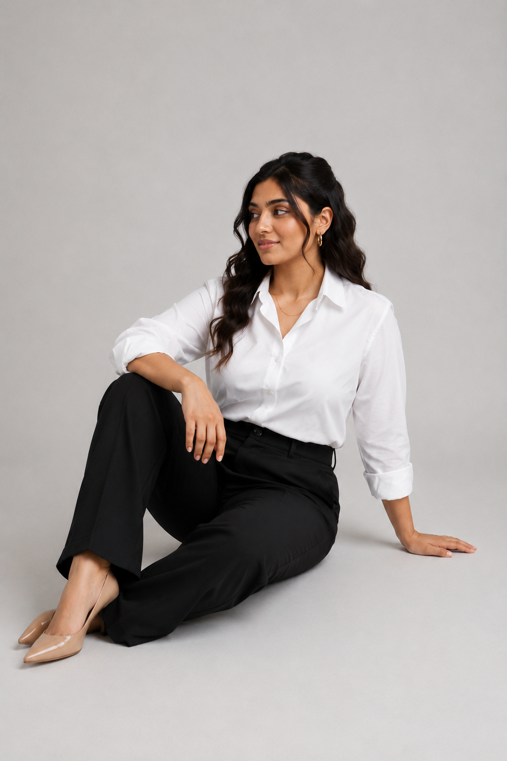 A woman sits on the floor against a light gray studio background, wearing a white button-up shirt by Purple Imli  with rolled sleeves and black trousers.