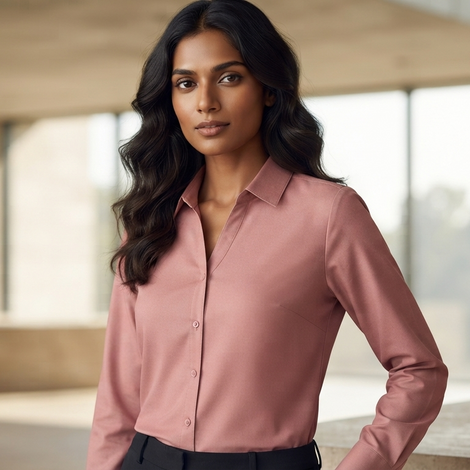 A woman with long, wavy dark hair wearing a dusty rose pink long-sleeve button-down shirt, dusty rose long-sleeve button-down shirt   by Purple Imli  standing in a modern, sunlit concrete building interior.