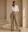 A woman stands in a minimalist beige architectural setting wearing a white button-down shirt by Purple Imli  and high-waisted beige wide-leg trousers, paired with dark brown heels.